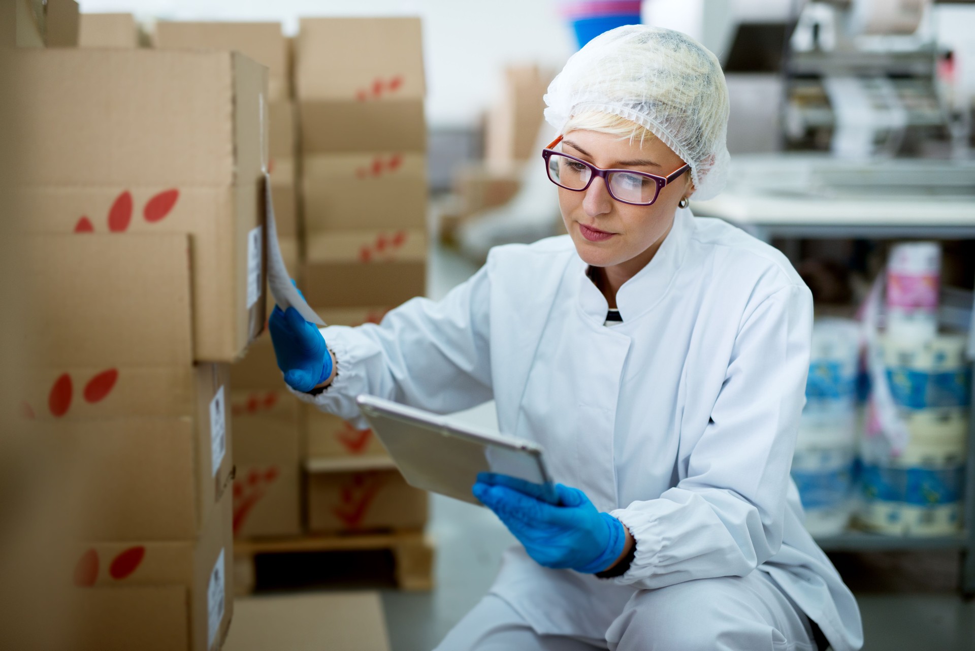 Young beautiful focused female worker in sterile cloths using a tablet to check correction of inventory in factory storage room.